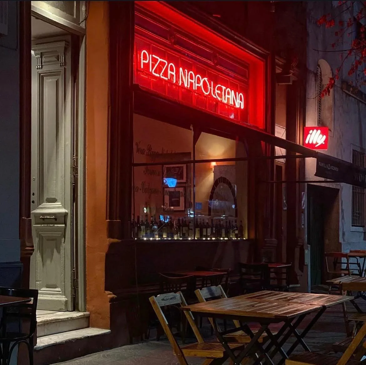Vidriera de Siamo nel Forno de noche con el cartel de neón 'Pizza Napoletana', botellas de vino y las mesas de madera en la vereda